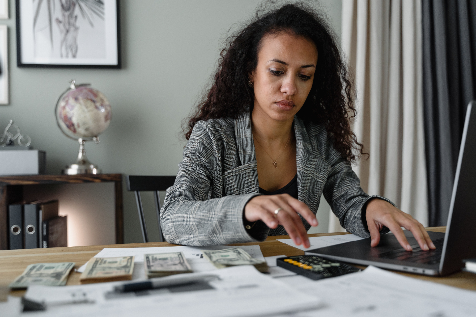 A woman sitting at a desk with her left hand on a laptop and her right hand on the phone's calculator. Next to her phone are dollar bills and in front of her are papers with notes. The woman is focused on calculating the cost of moving, including the security deposit deductions.