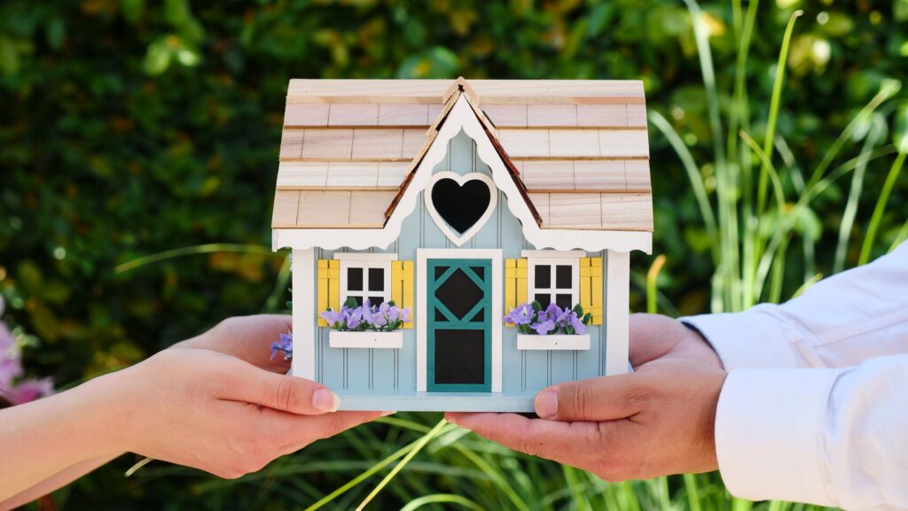 A miniature house model is being handed to someone else in an outdoor landscape. The house model is painted blue with two windows painted yellow decorated with purple flowers. There is also an overhead heart-shaped window on the model. This image symbolizes the joy of buying your first investment property.