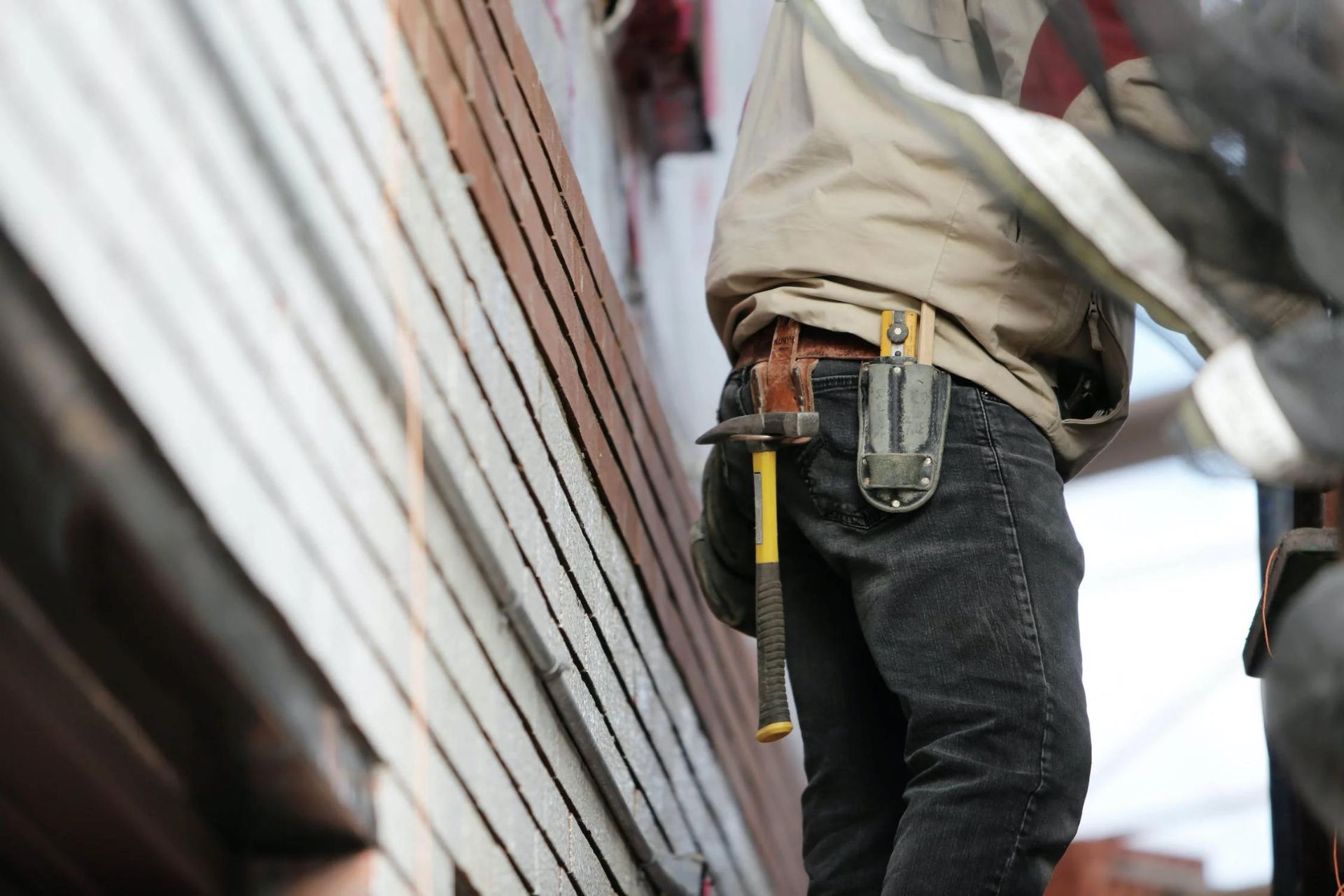 A person in a tool belt with a hammer and level tool, inspecting the house exterior for property preventative maintenance.