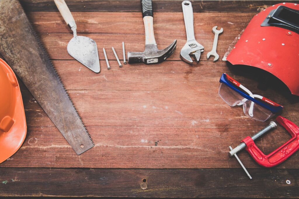 A selection of essential tools, including an orange safety helmet, saw, trowel, nails, hammer, wrench, screw, safety goggles, and a red clamp, thoughtfully organized on a sturdy wooden table—a perfect setup for handling a tenant maintenance request.