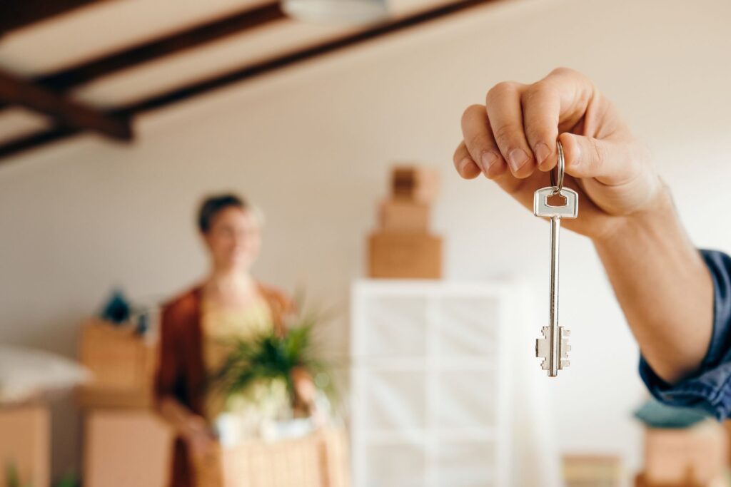 A hand holds the key to a vacant rental property, with a woman in the background holding a box. Behind her, stacks of boxes suggest a recent move-in, emphasizing the transition into the newly leased property.