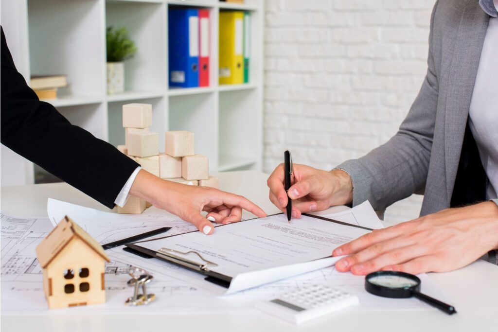 In a grey office, two individuals in black and grey suits stand around a white desk emphasizing only their hands. On the desk, a miniature house model, keys, a magnifying glass, and an Orlando lease agreement are featured, with one person pointing and the other signing.