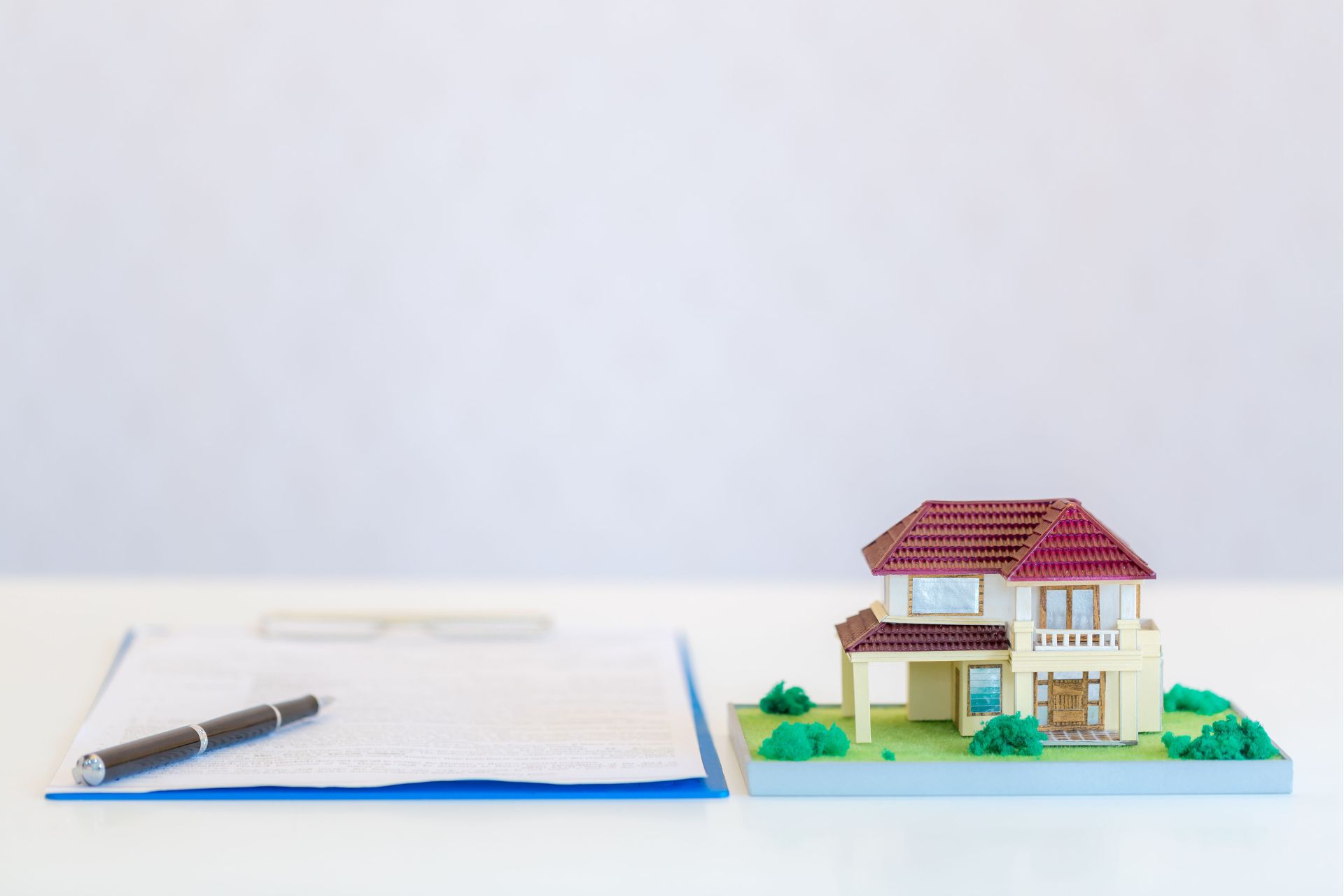 A pen rests atop a document on a blue clipboard, with a miniature house model featuring a red roof and a green yard placed to the right, symbolizing the concept of a 1031 exchange in real estate investment.